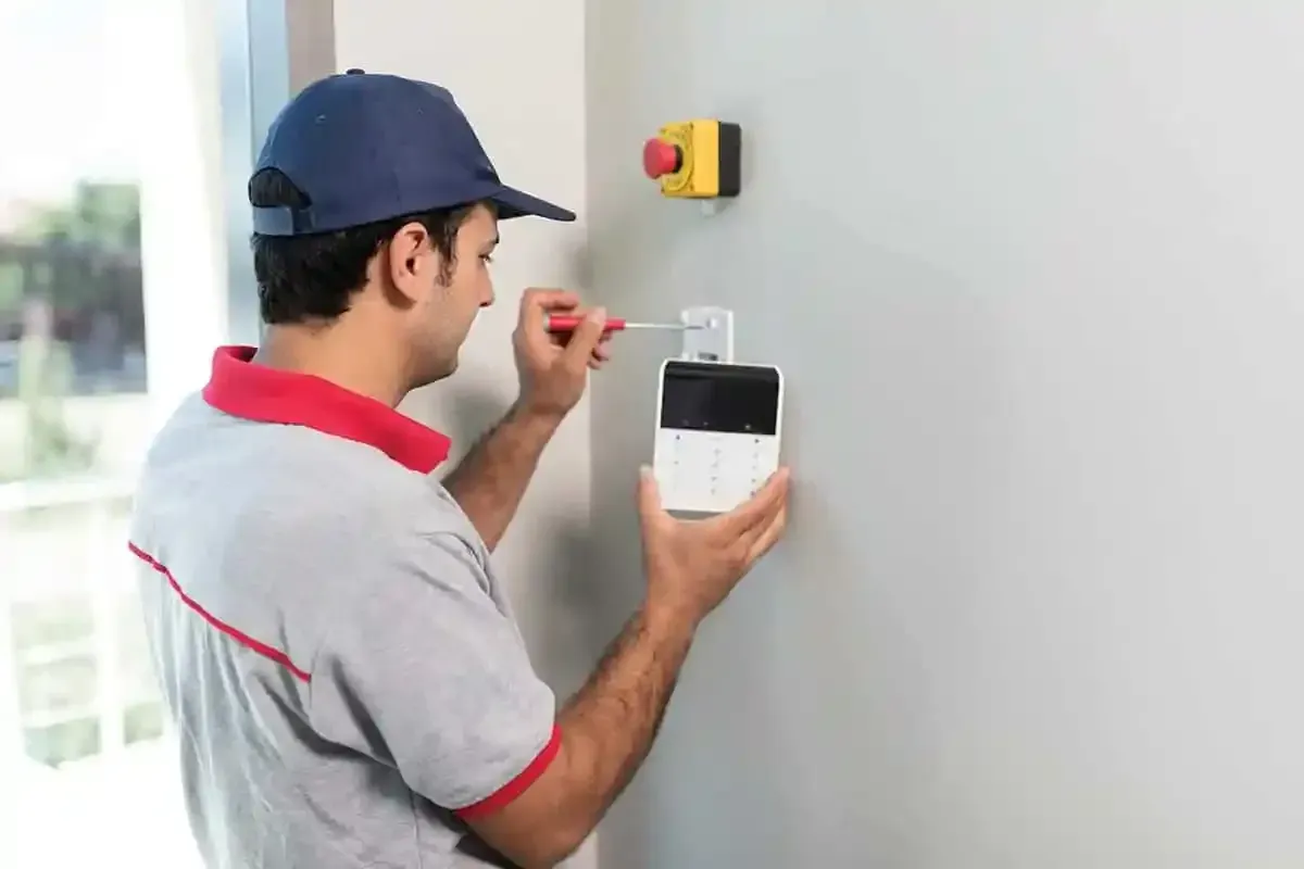 Man in uniform installing a security device on a wall near a red button.