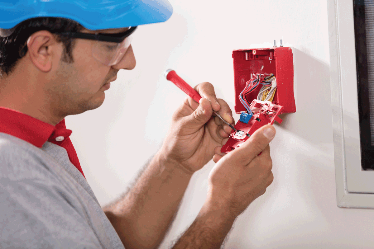 Electrician working on electrical box, wearing a hard hat and safety glasses, inside a building.