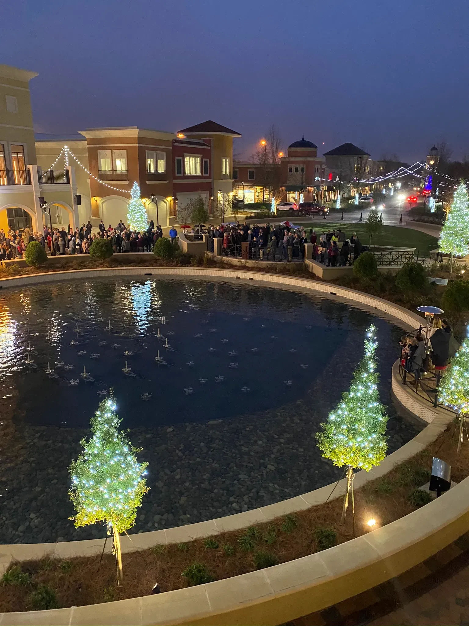 Evening shot of a town square with holiday lights and trees around a fountain; crowd gathers.