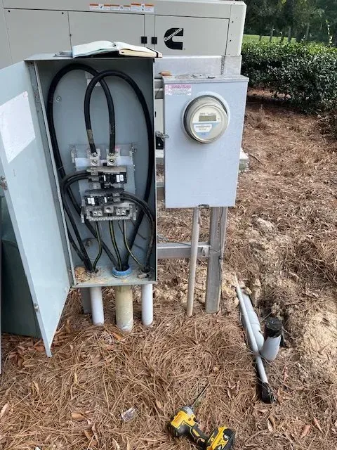 Electrical box with cables and meter; ground with tools and generator in background.