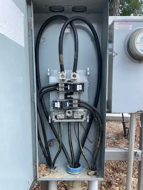 Open electrical panel with black wires connected to breakers, mounted outside near a meter.