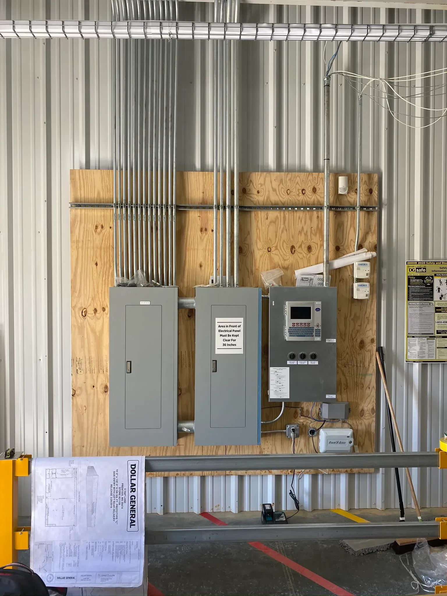 Electrical panel installation on plywood backing in a metal-walled industrial setting. Conduit runs overhead.