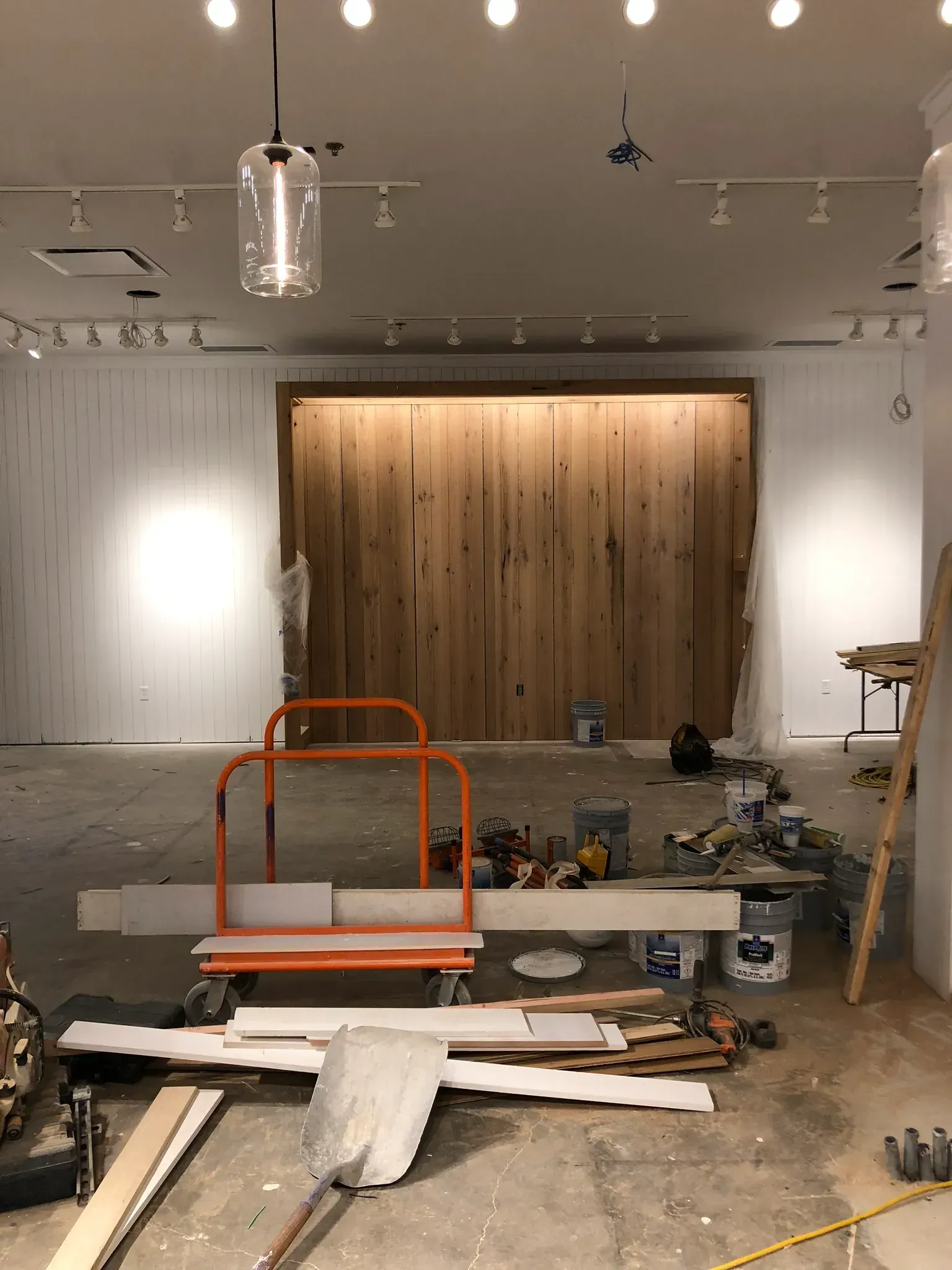 Construction site with wooden wall, cart, and tools; unfinished white walls and ceiling; overhead lighting.
