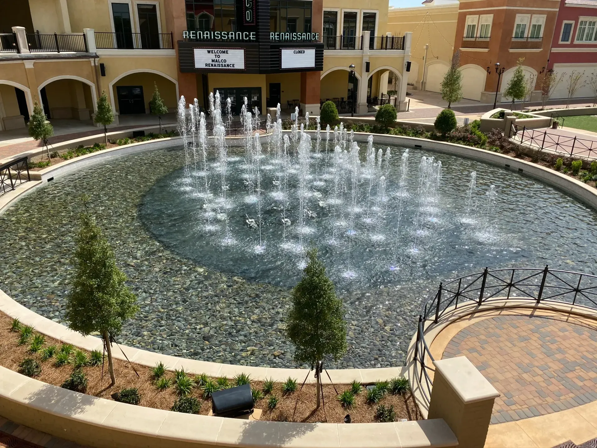 Circular fountain in a courtyard, spraying water; surrounded by trees, buildings, and a brick patio.