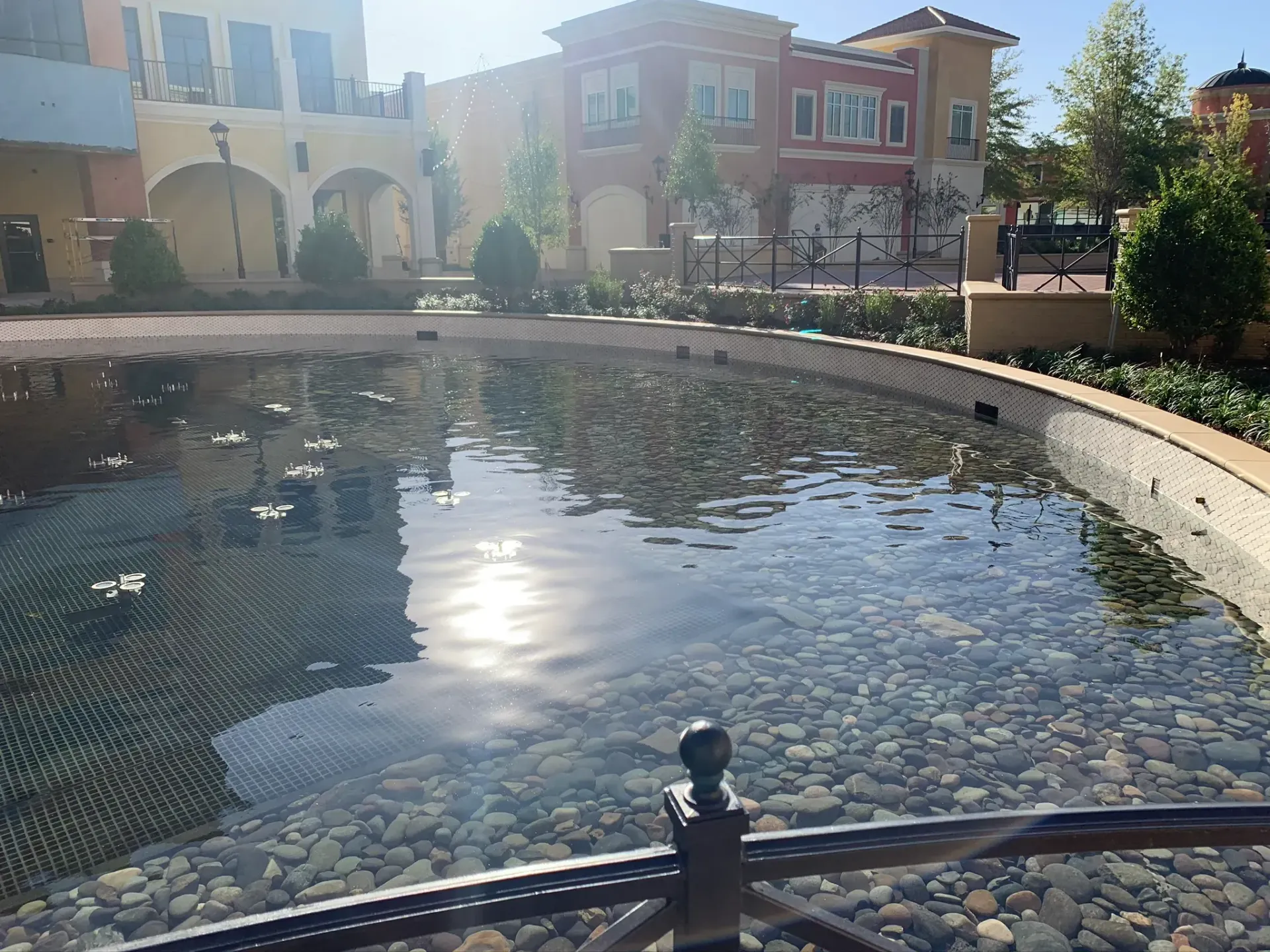 Pond with cobblestone bottom reflecting sunlight, in front of colorful buildings.