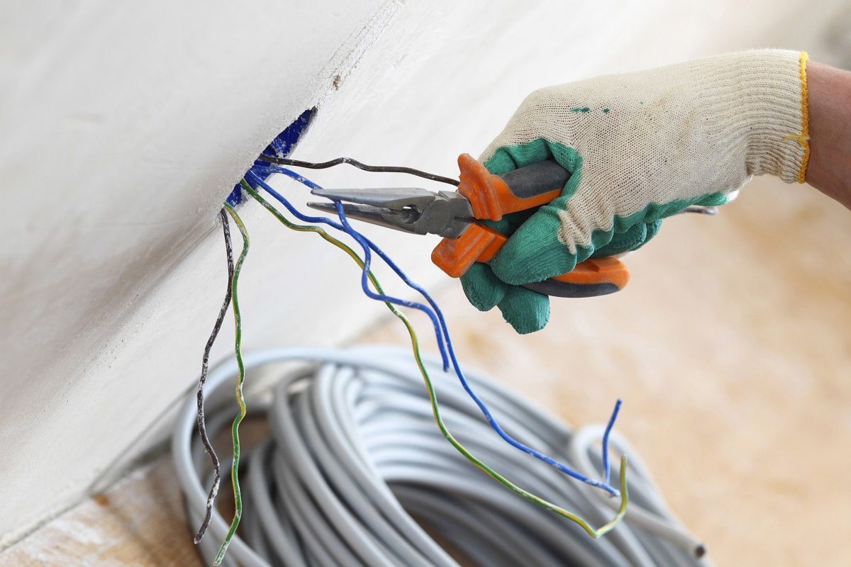 Gloved hand using pliers on electrical wires emerging from wall. Wires are blue, green, and black.