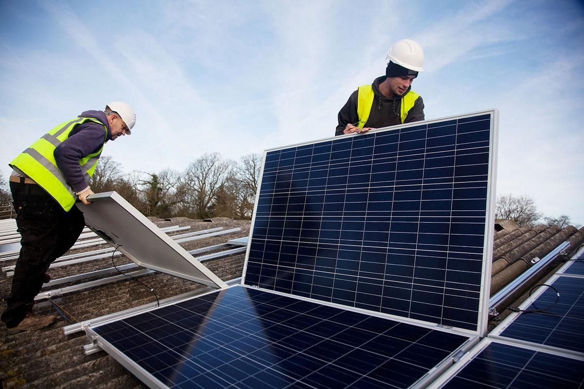 Two workers installing solar panels on a rooftop. They wear safety vests and helmets. Blue sky in the background.