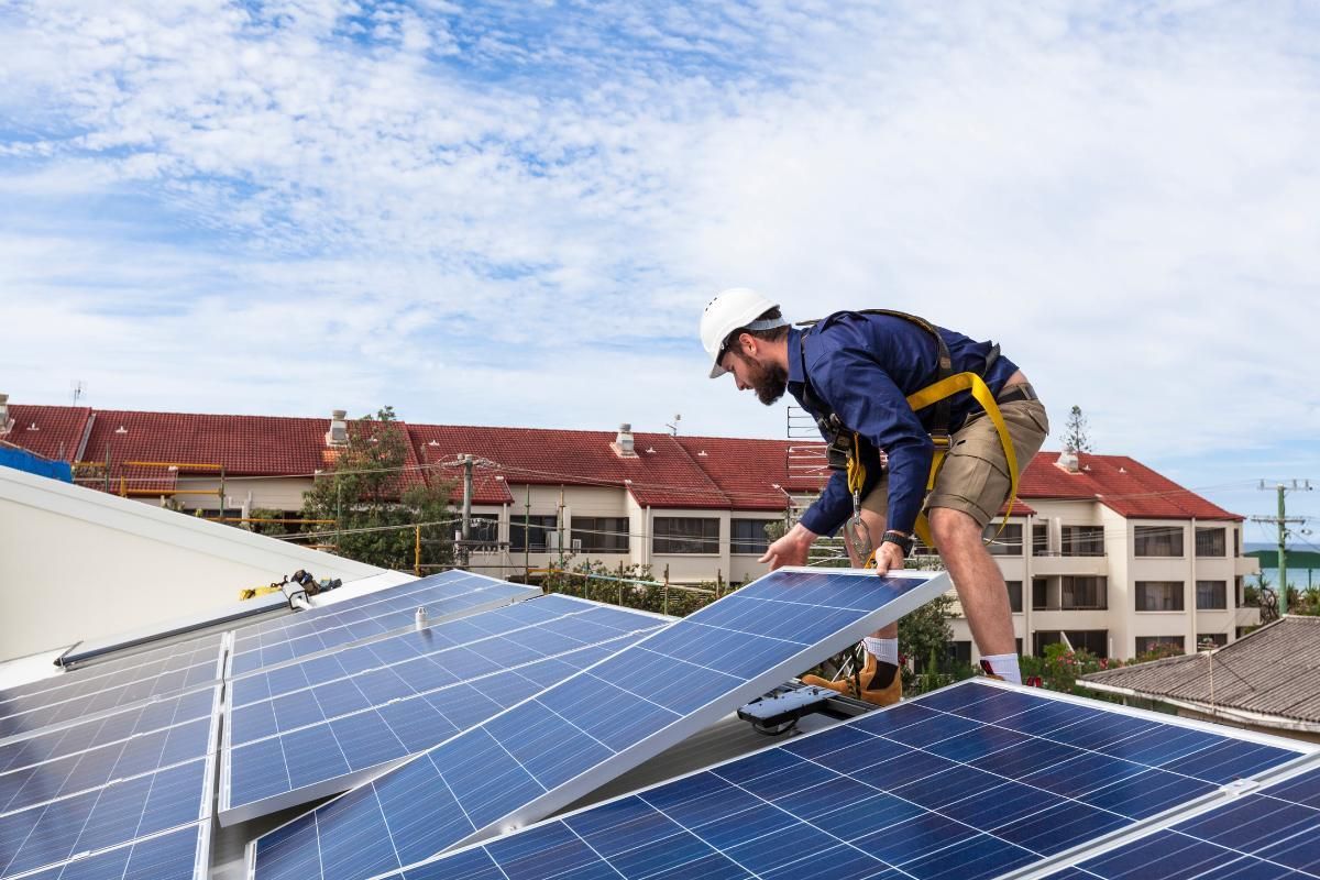 Man in safety harness installs solar panel on a rooftop with buildings and blue sky in the background.
