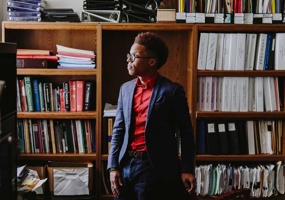 Person in suit and glasses stands by bookshelf, looking to the side. Red shirt, navy jacket, books visible.