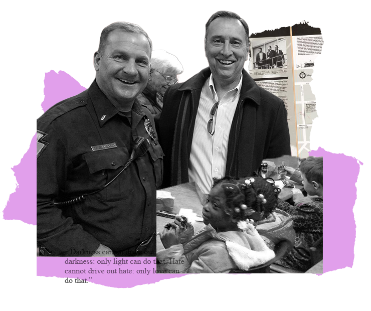 Two men smiling, one in a uniform.  They stand near a table with objects.