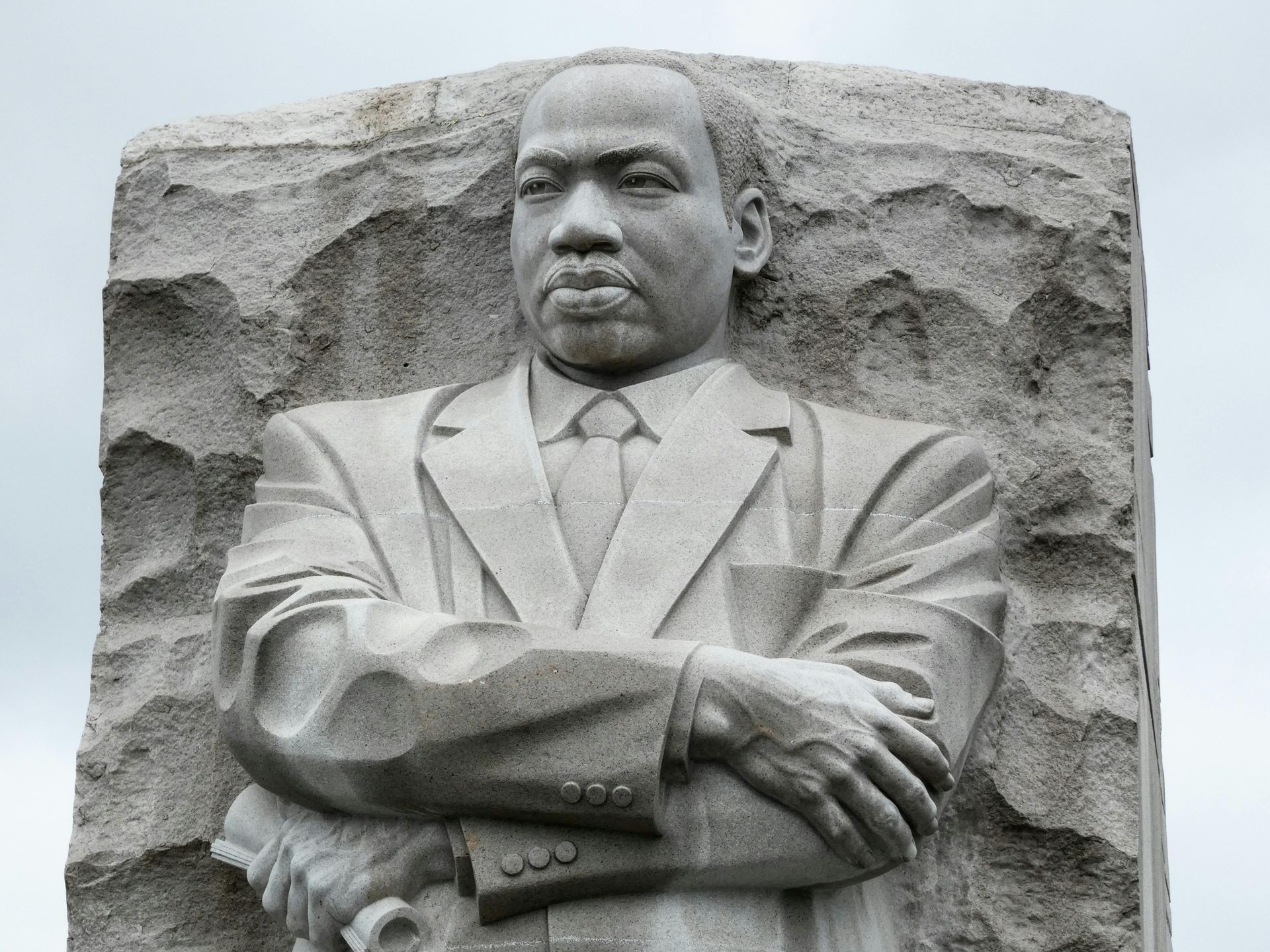 Stone carving of Martin Luther King Jr. with arms crossed, set against a rough stone backdrop.