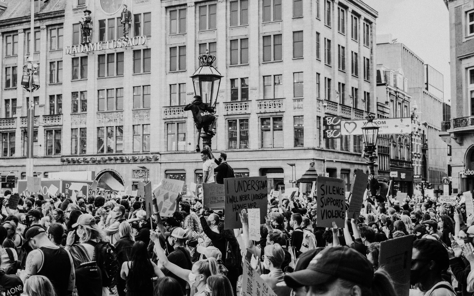 Large crowd protesting in a city square, holding signs. Buildings in the background.