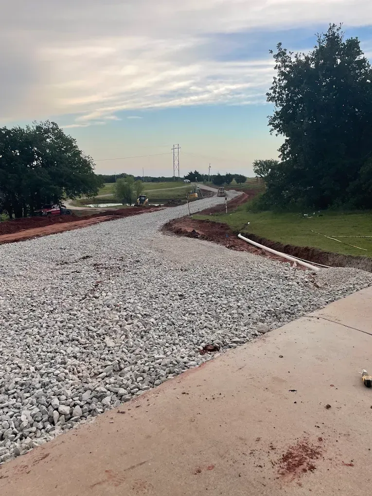 Gravel road under construction curves toward a horizon with trees, blue sky, and a light pole.
