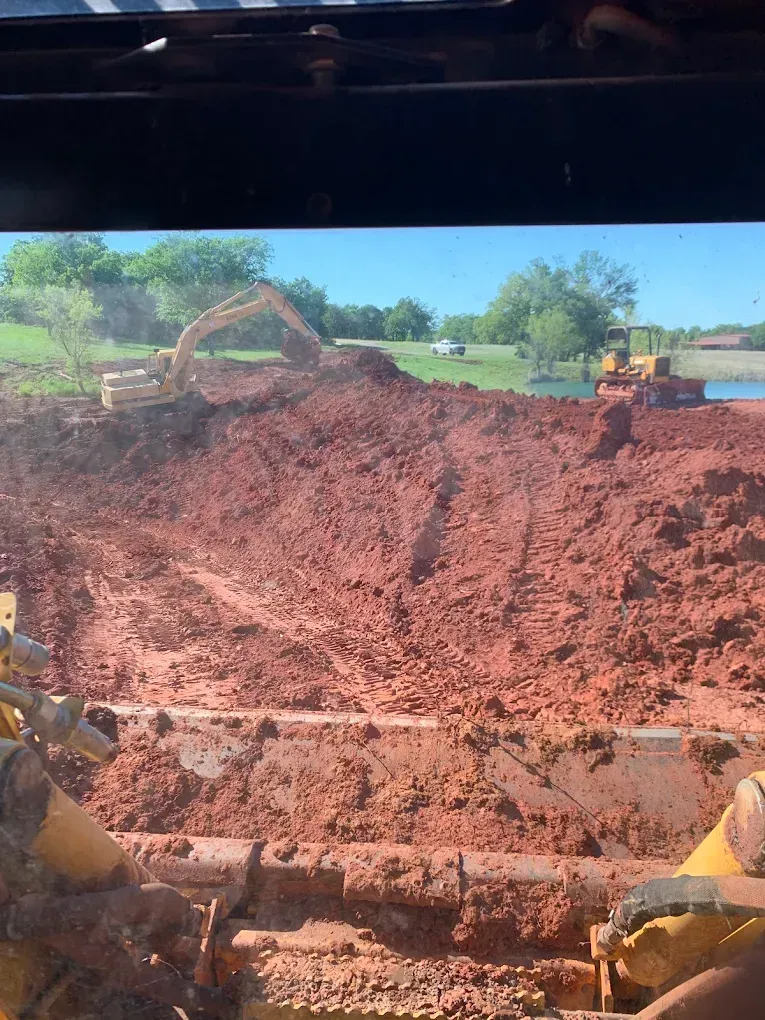 View from a heavy machine shows two excavators moving red soil at a construction site near water and trees.
