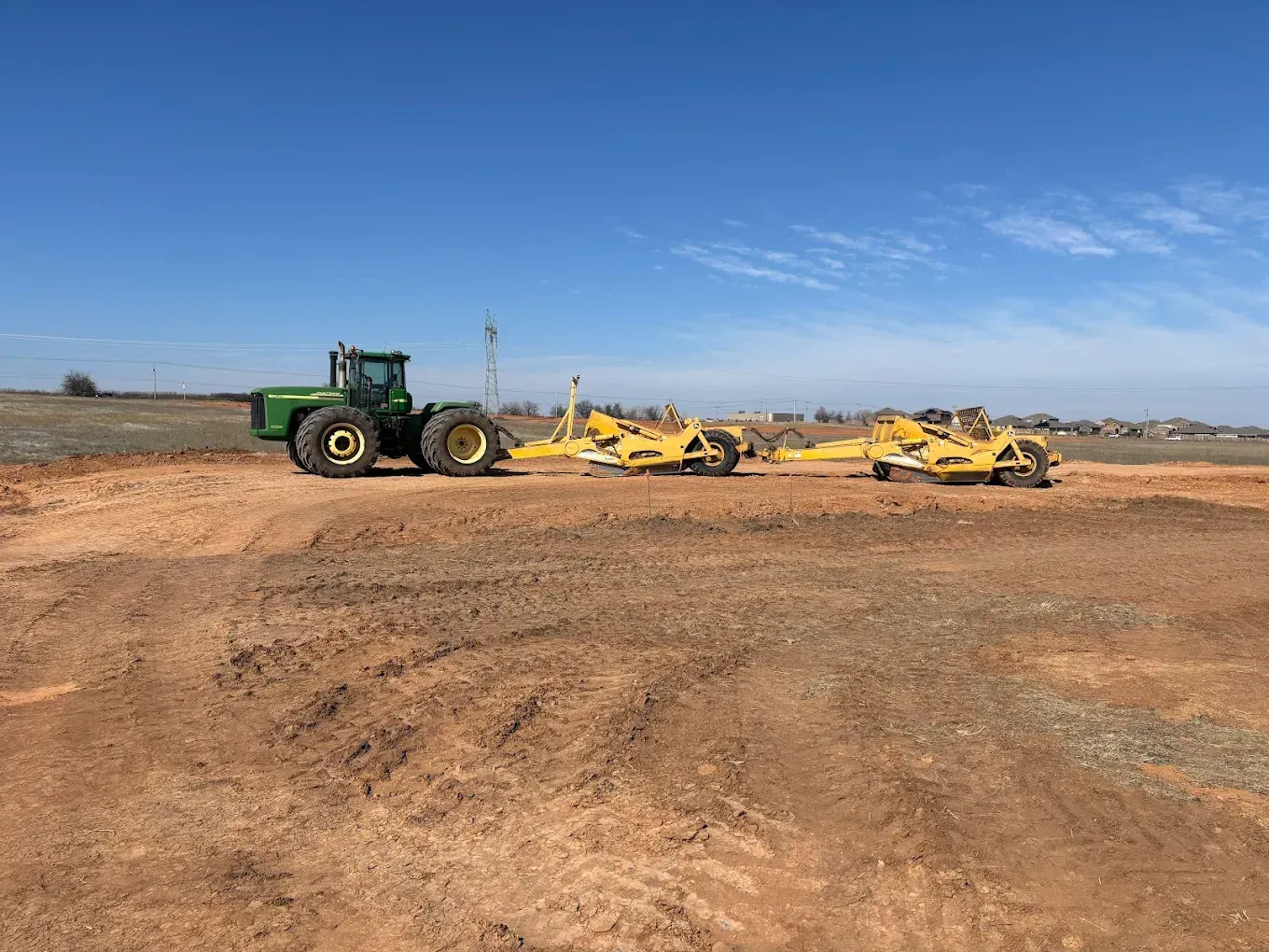 Green tractor pulling yellow grading equipment on a dirt field under a blue sky.