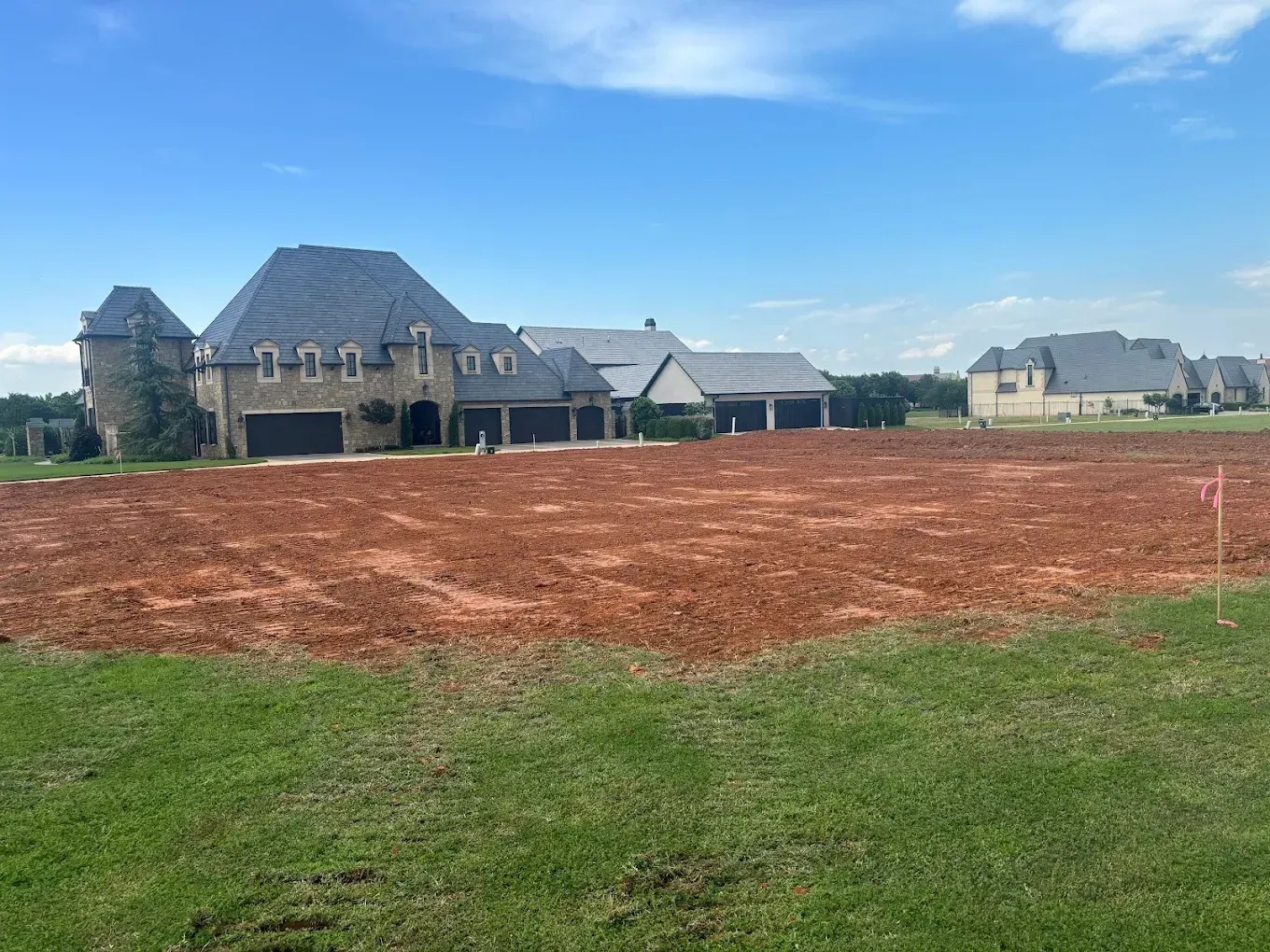 Cleared lot in front of large houses with dark roofs, under a blue sky.