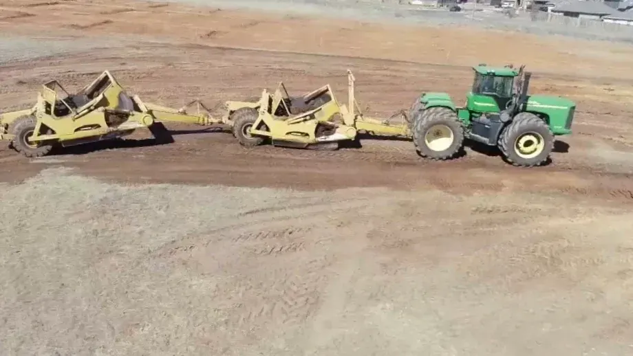 Green tractor pulling two yellow scrapers on a dirt field.