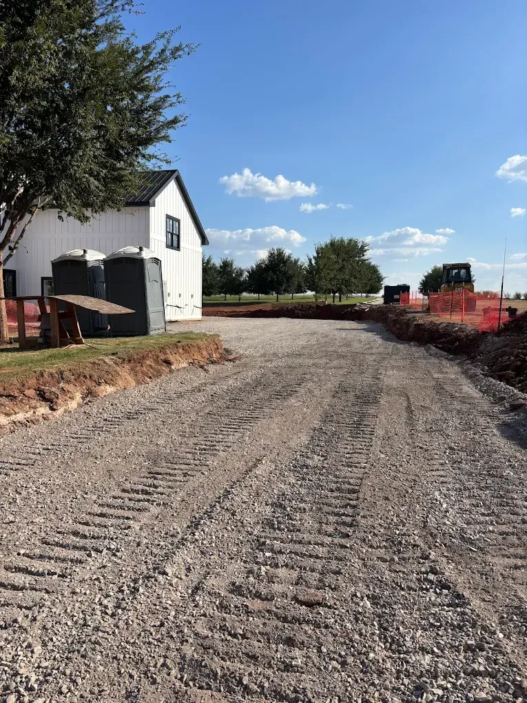 Gravel driveway leading to a white building under a blue sky. Construction equipment and portable toilets are present.