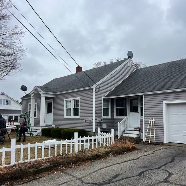 Gray house with white picket fence, driveway, garage, and overcast sky.