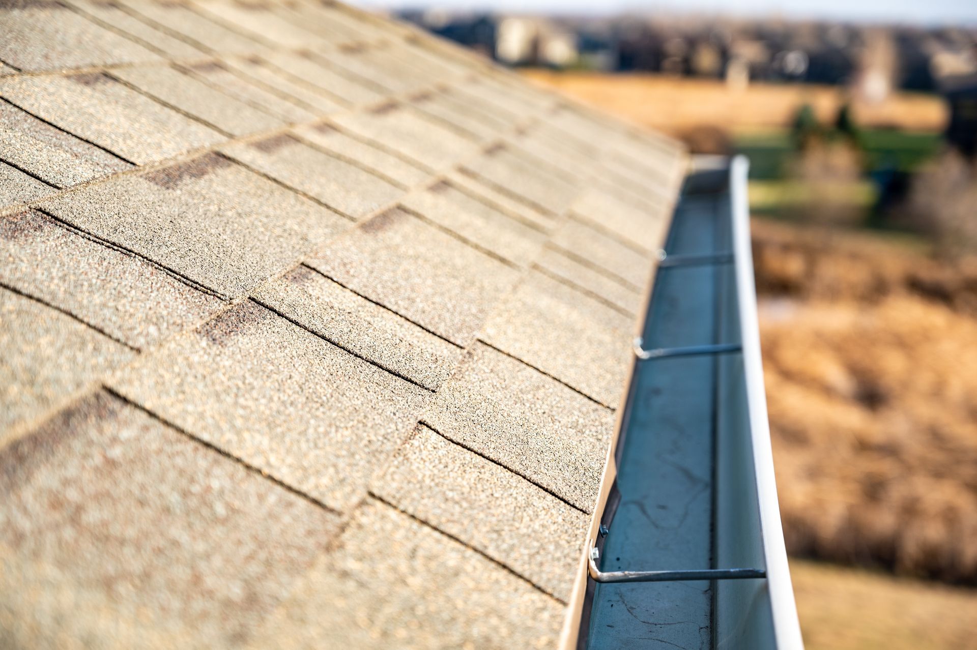 A close-up, high-angle view of a beige shingled roof edge with a metal gutter attached.
