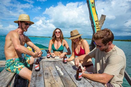 Friends in straw hats relax at a wooden picnic table on a sunny beach with drinks and surfboards.
