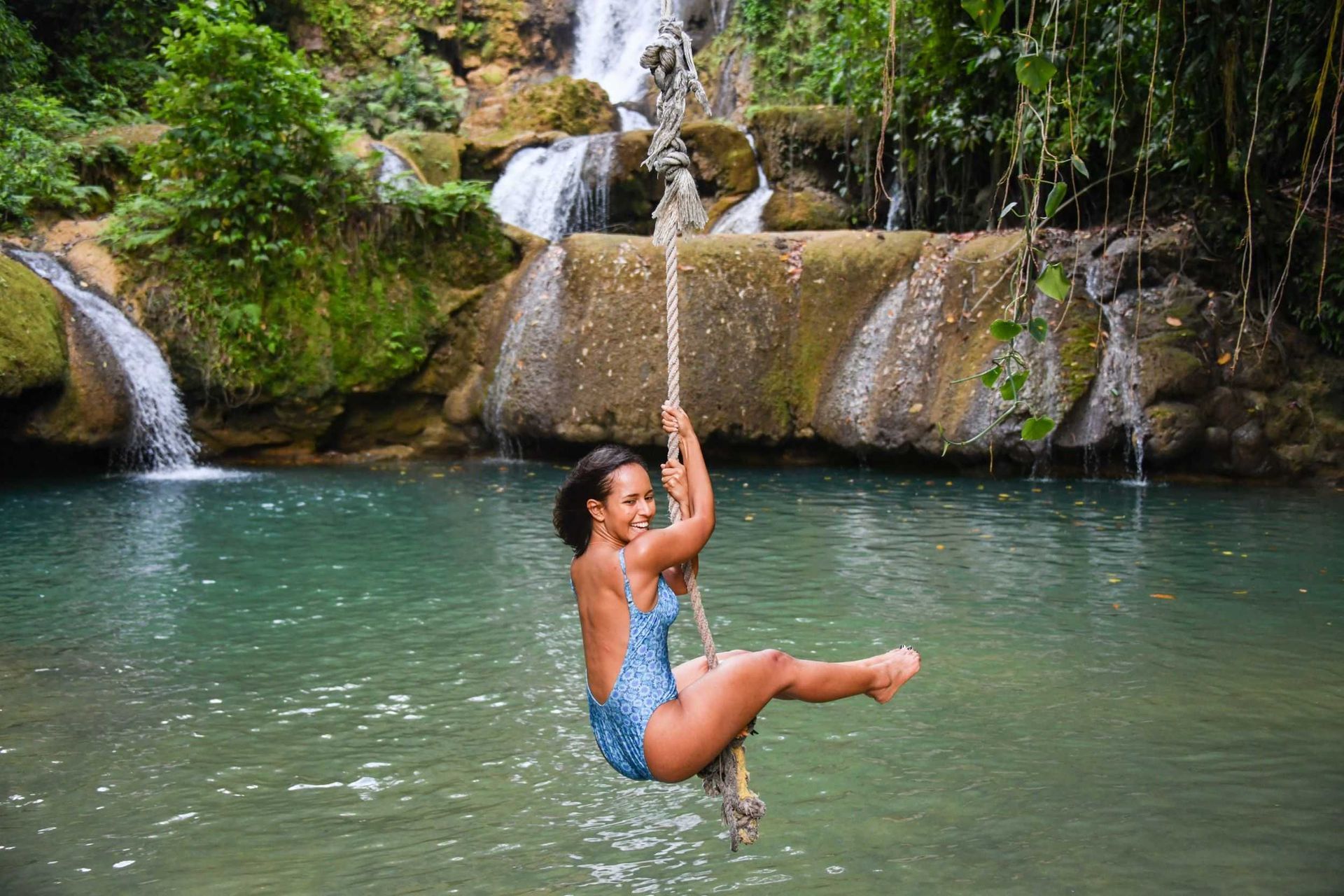 Person in a blue swimsuit swinging on a rope over a turquoise waterfall pool