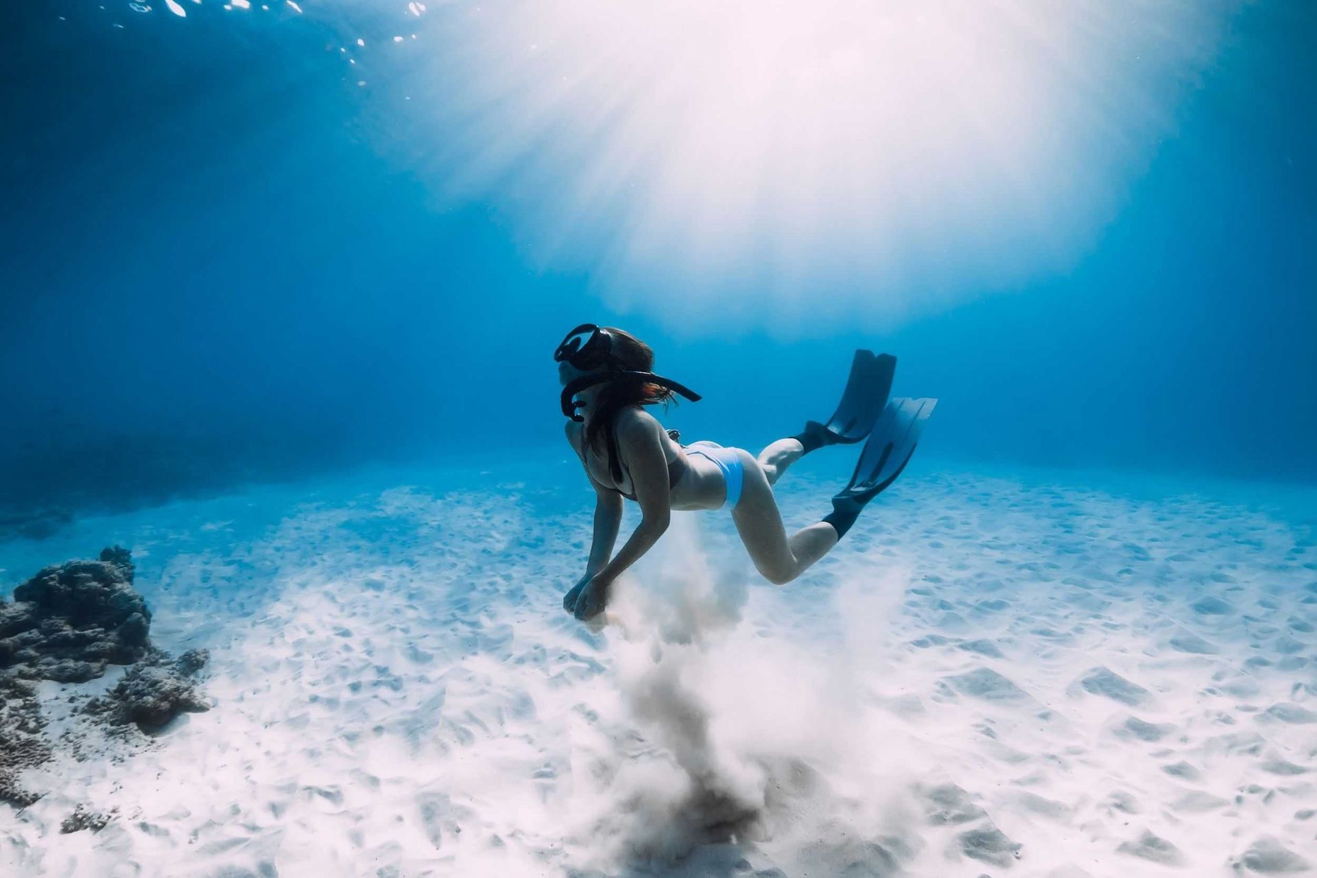 Two divers underwater above sandy seabed, with bright sunbeams filtering through blue water.