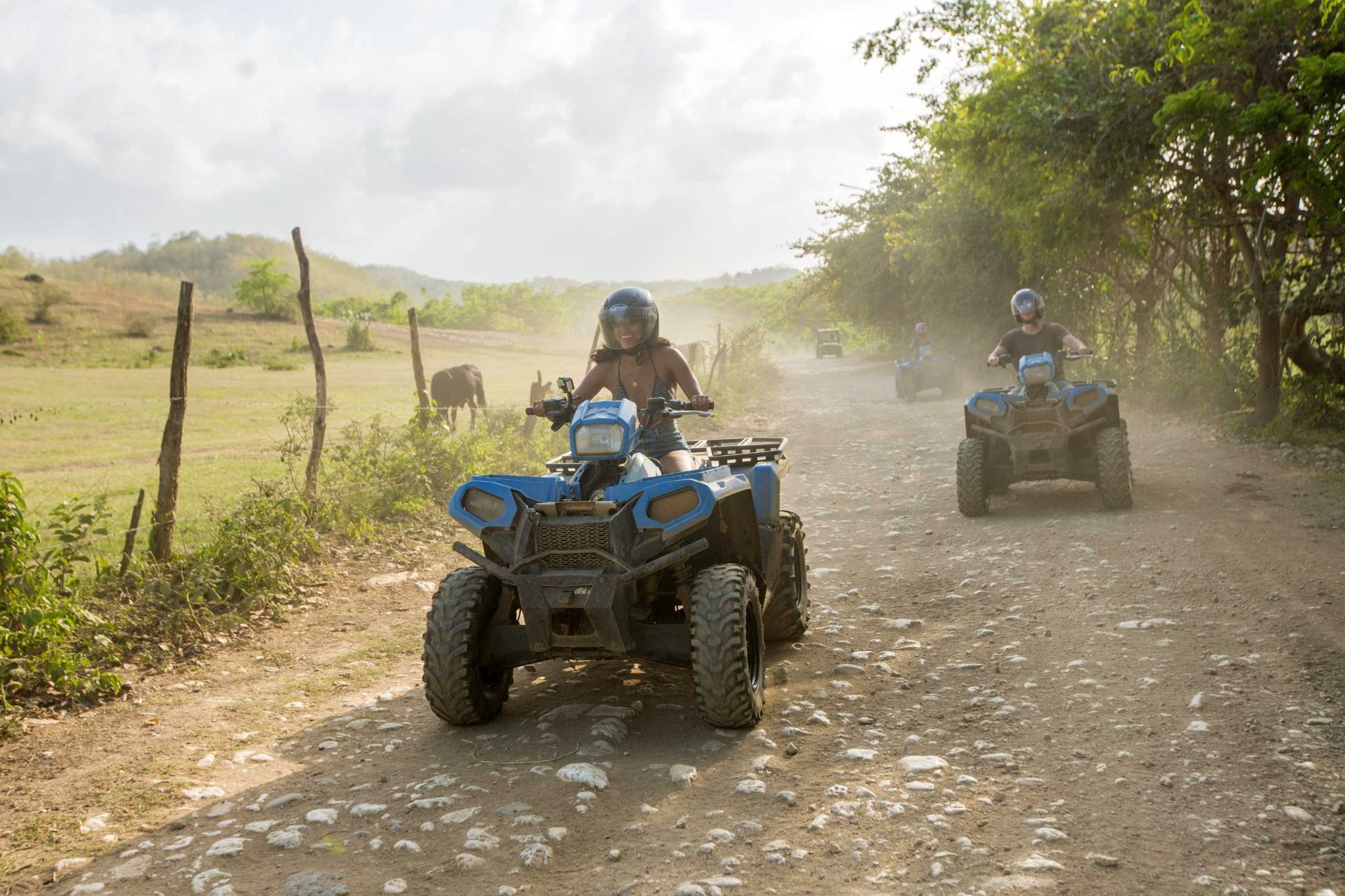 Two riders on blue ATVs driving along a dusty rocky road through a green rural landscape
