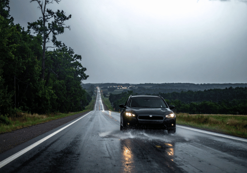 Car driving through rain on a wet road with headlights on and water splashing beneath tires.