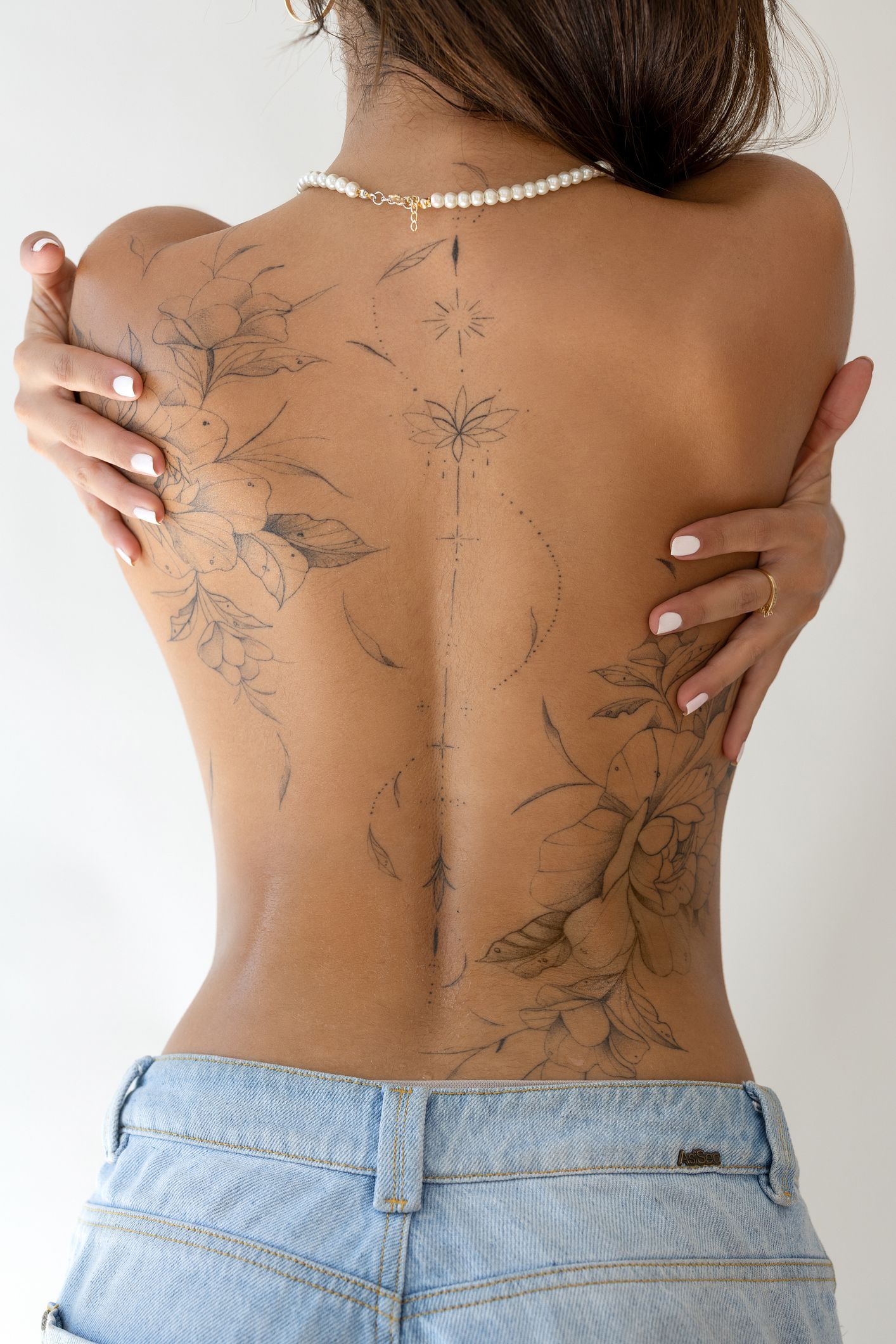 Woman's bare back with dark, floral-like patterns. She embraces herself, wearing a pearl necklace and jeans.