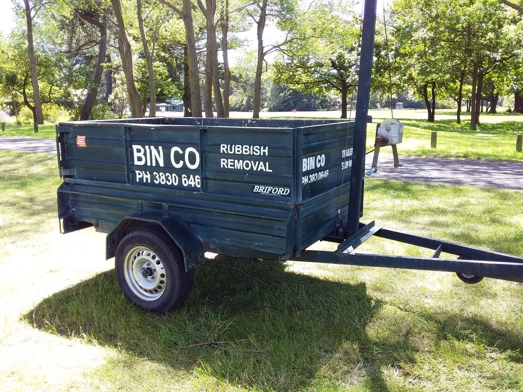A dark green rubbish removal trailer parked on a grassy field with trees in the background.