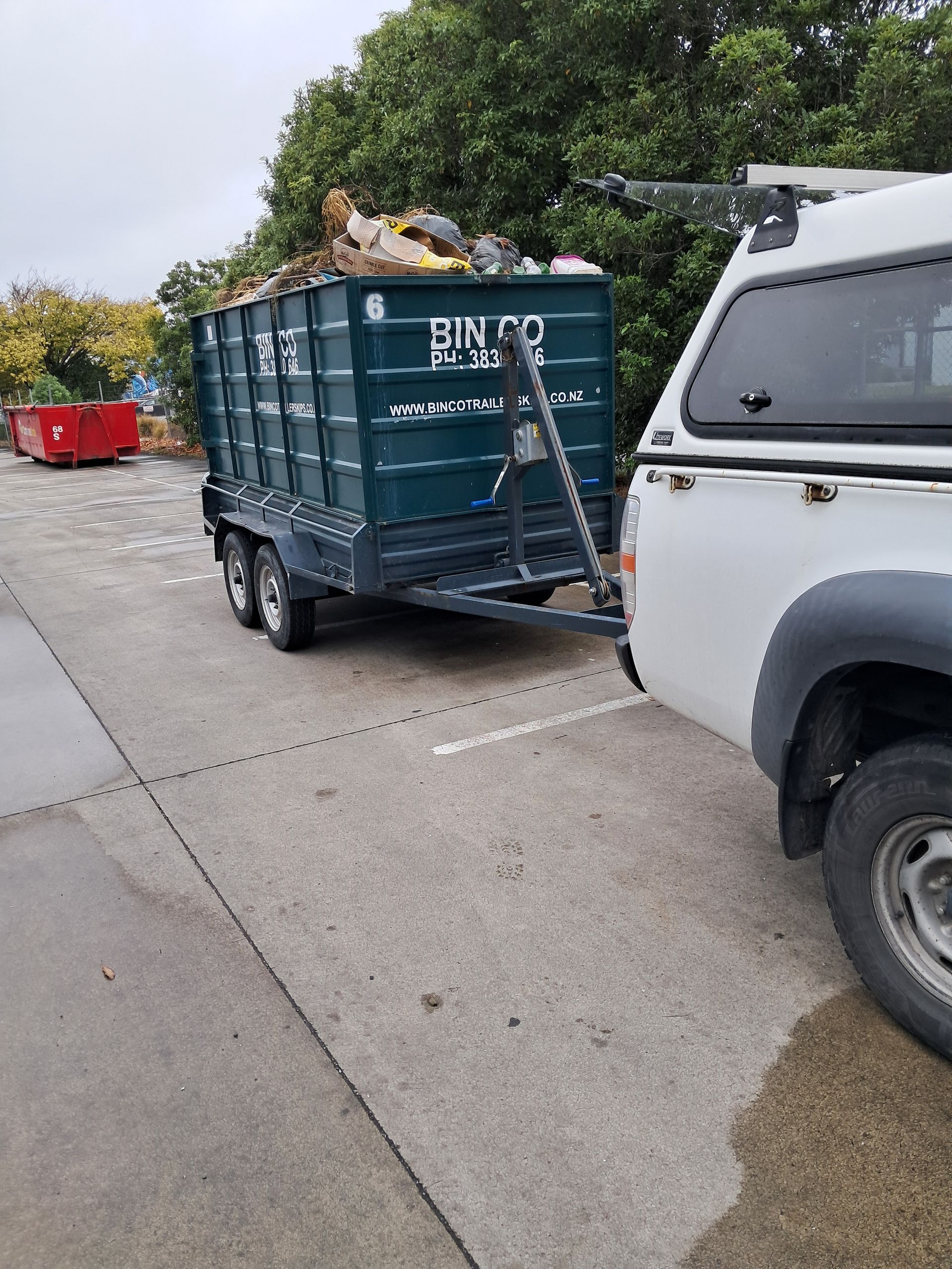 A white pickup truck hitched to a dark green waste bin trailer parked on a concrete lot.