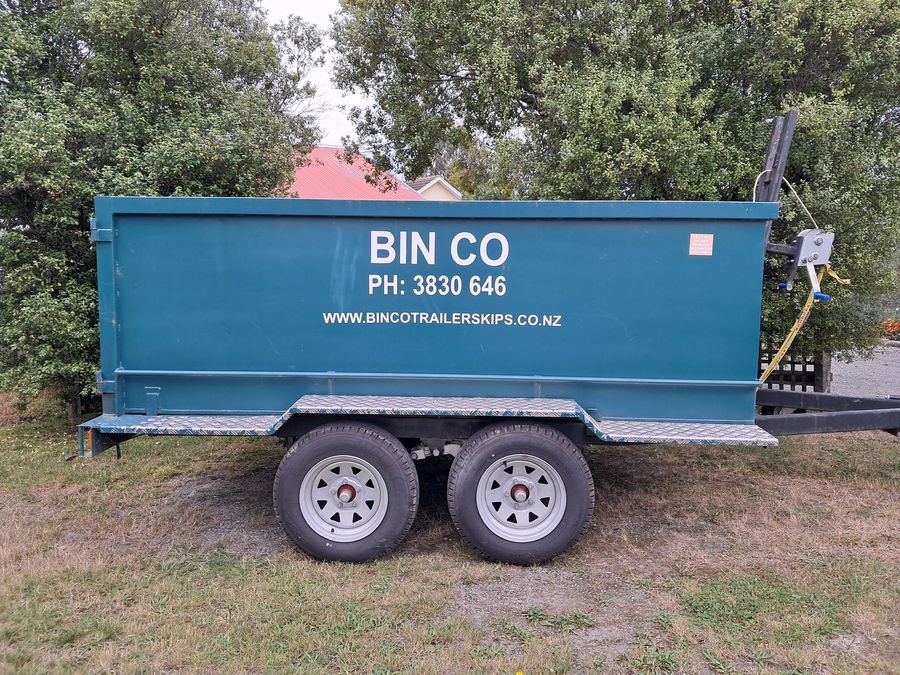 A teal Bin Co skip trailer parked on grass in front of trees.