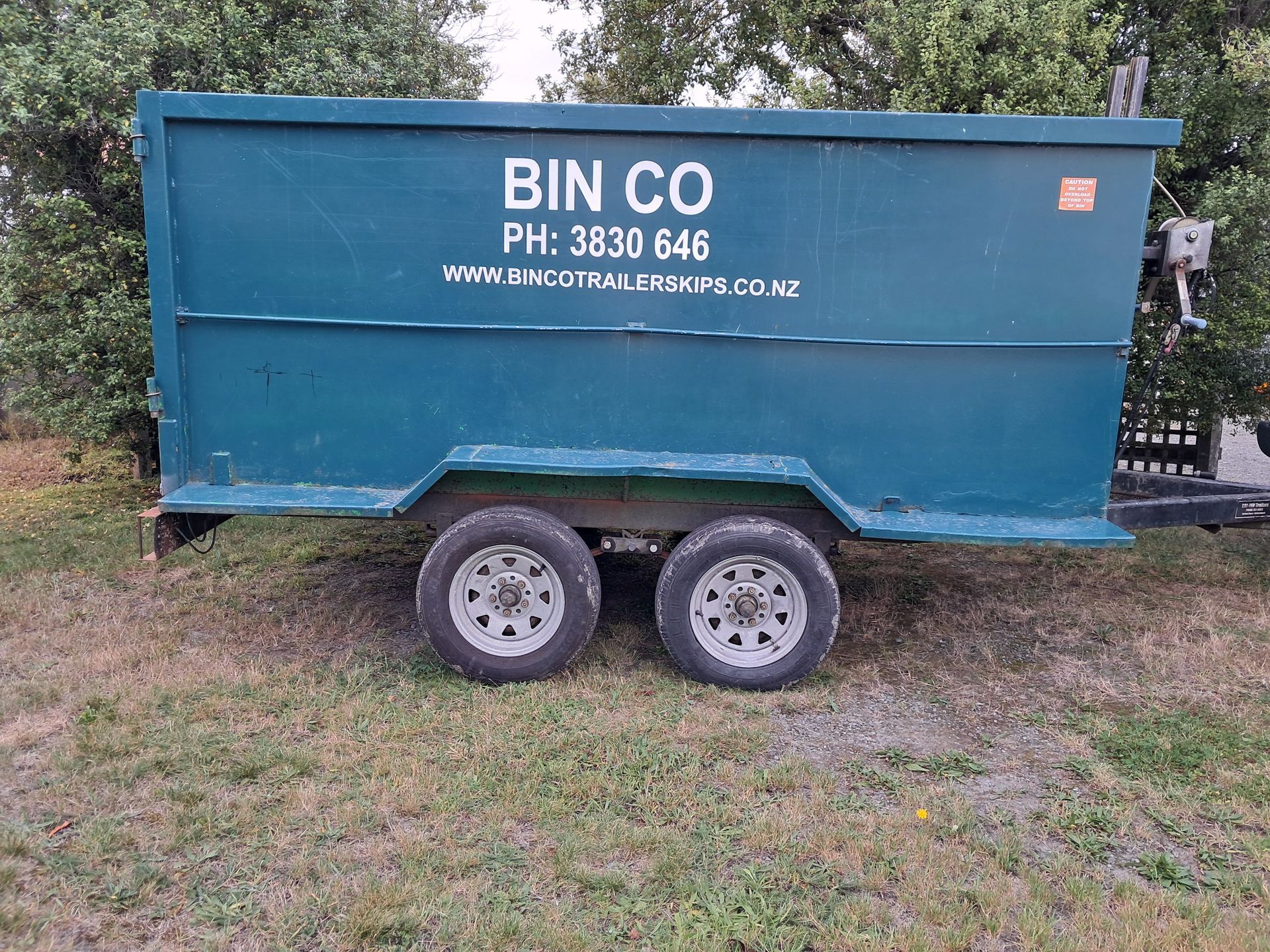 A teal, dual-axle skip bin trailer parked on grass, labeled BIN CO with a phone number and website address.