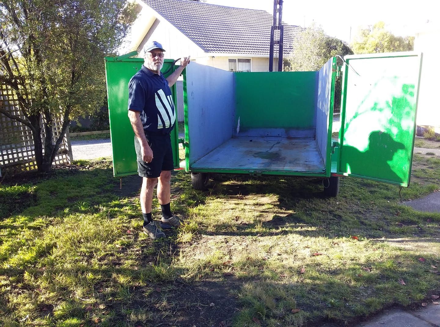 A person standing next to an open, empty green dump trailer in a grassy yard on a sunny day.