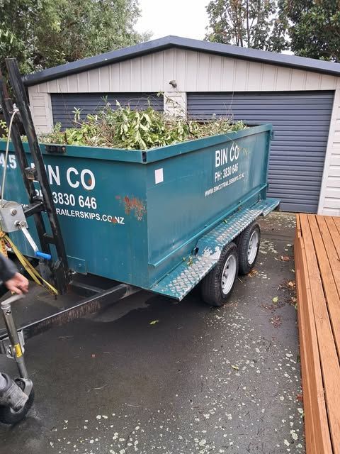 A teal waste disposal trailer filled with garden debris is parked in a driveway in front of a garage.