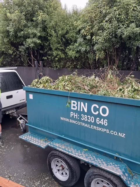 A teal Bin Co trailer skip filled with green garden waste, hitched to a white vehicle parked in front of trees.