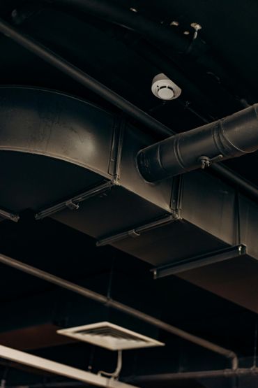 Black industrial ceiling with ducts, pipes, and a security camera.