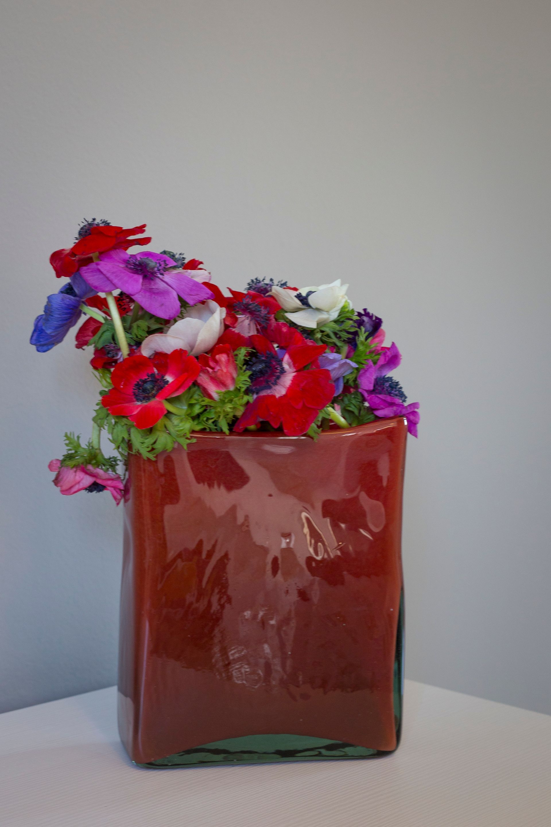 Fiori nelle tonalità del rosso, viola e bianco in un vaso rettangolare rosso-marrone contro una parete bianca.