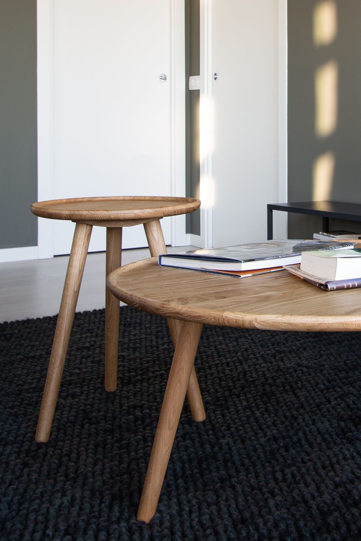 Due tavoli in legno chiaro con tre gambe angolate su un tappeto scuro. Sfondo: porte bianche, parete grigia.