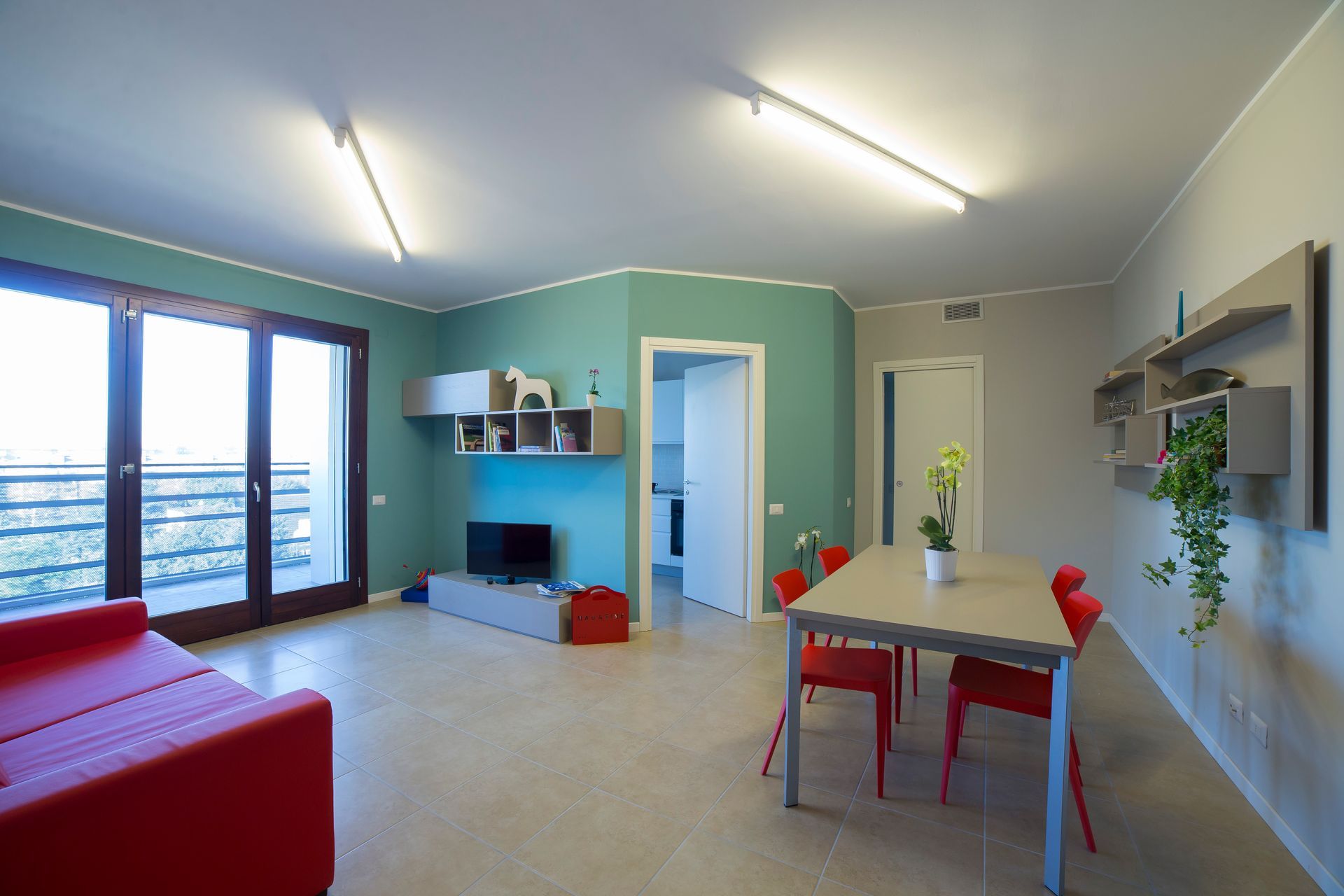 Ampio soggiorno con divano rosso, tavolo da pranzo, parete turchese, porte-finestre e scaffali aperti.