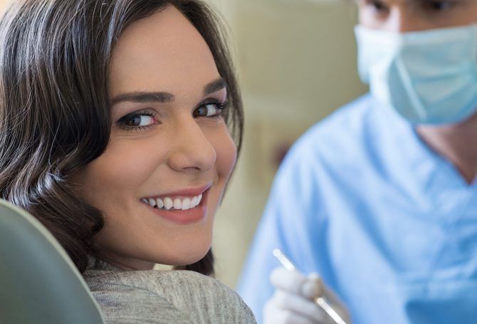 A smiling person in a dental chair looks toward a dentist wearing a blue scrub top and a face mask.