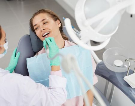 A dentist in a white coat and green gloves performs a dental examination on a patient sitting in a reclining chair.