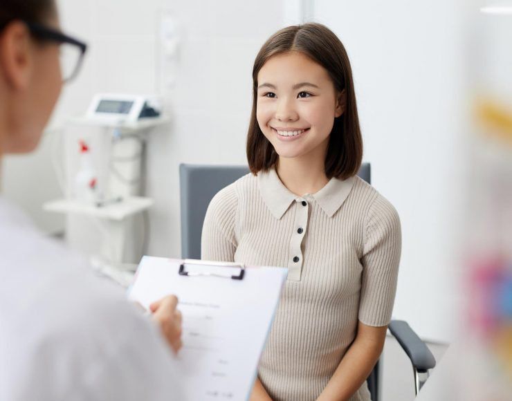 A person in a white coat holds a clipboard while consulting with a smiling individual in a medical office.