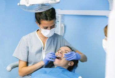 A dentist in scrubs and a mask performs a dental exam on a patient in a light blue room.