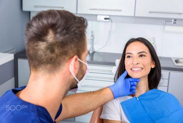 A dentist in blue scrubs and a mask examines a smiling patient’s jaw in a clinical dental office.