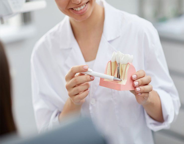 A dentist in a white coat uses a pen to point to a dental implant model, explaining the procedure to a patient.