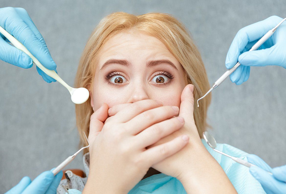 Woman with panicked expression, hands covering mouth, surrounded by dental tools held by gloved hands.