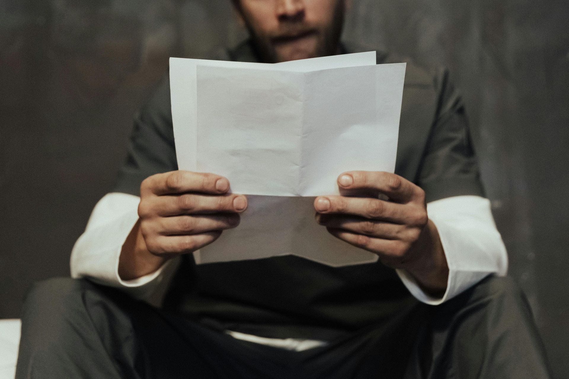 Person in a gray uniform reading a folded paper; seated, focused expression. Dark, grainy background.