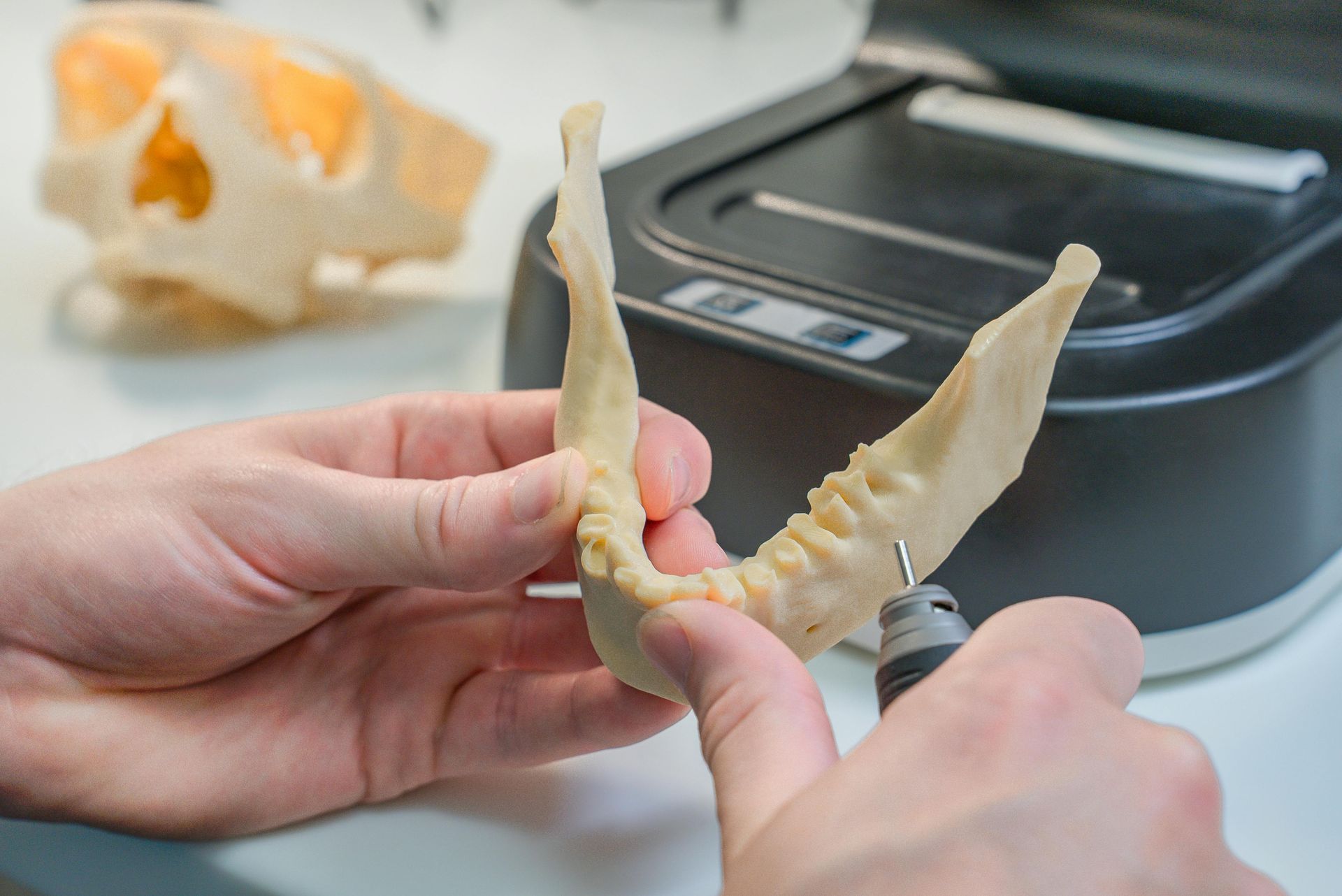 Hands holding a model jaw, using a small drill. A skull and device are in the background.